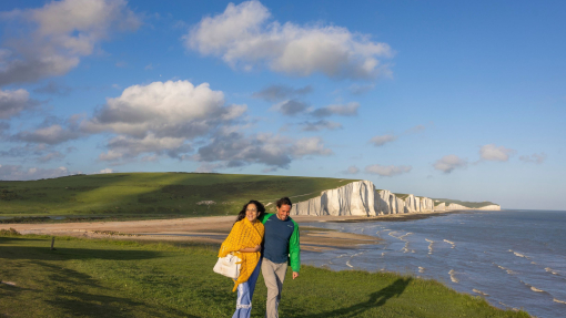 A couple walk together on a headland with white chalk cliffs in the background.