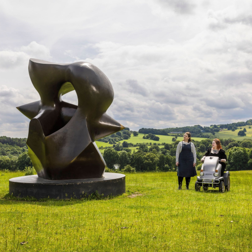 Two women look at a large sculpture set in green gardens with landscape beyond