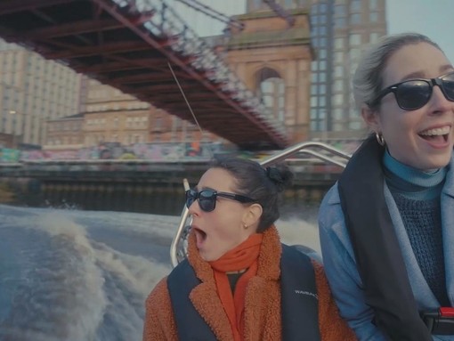 Two women on a motorboat on a river, with a bridge and buildings in the background