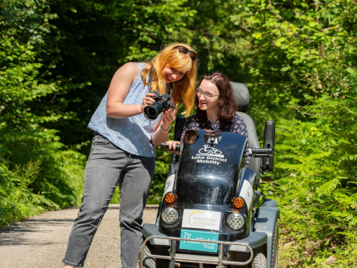 A woman stands holding a camera showing the pictures to a woman who sits in a tramper