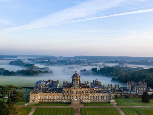 Large country house seen from above with formal gardens and wooded countryside behind covered in low lying cloud