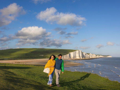 A couple walk together on a headland with white chalk cliffs in the background.