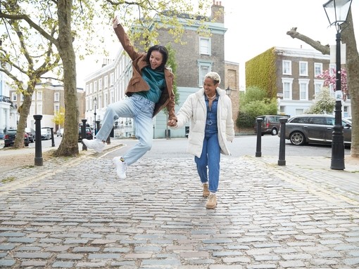 Happy young woman jumping while holding girlfriend's hand walking on a footpath in London street
