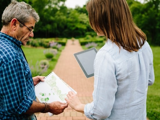 Rear view of male gardener sharing garden plans with female garden designer holding an iPad stood on a path in a garden