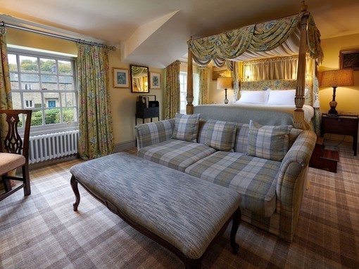 Bedroom at the Devonshire Arms at Pilsley, Derbyshire with a grey tartan upholstered sofa and large foot stool in the foreground, matching tartan carpet and floral curtains and matching draped four-poster bed in the background.