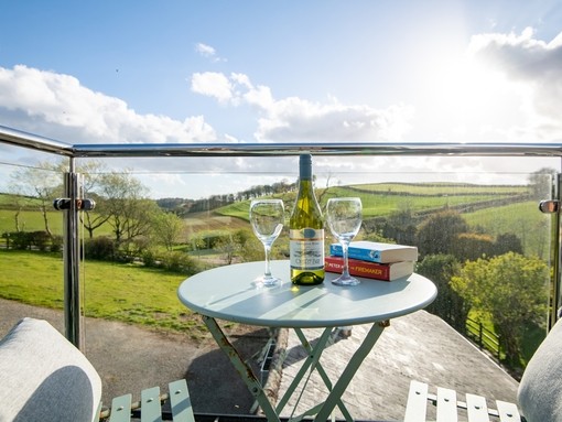 View from the balcony at Pitt Farm Holiday Cottage, Devon with a bistro table and chairs set with two novels, tw wine glasses and a bottle of wine with a rural landscape in the background. Bronze winner of the Self Catering Accommodation of the Year in the VisitEngland Awards for Excellence 2022