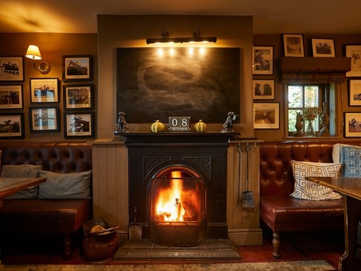 A fireplace with a lit fire flanked by two leather sofas with tables. The wall is covered in numerous framed artworks. The Duncombe Arms - Bronze award winner for the Pub of the Year at the VisitEngland Awards for Excellence 2023.