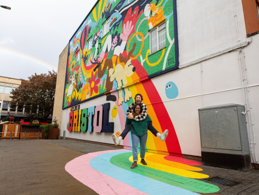Two people playfully smiling and posing in front of a colourful mural.