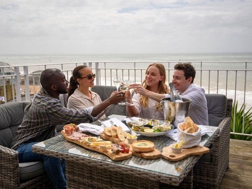 Two men and two women laugh together while sat on a hotel balcony with the sea beyond