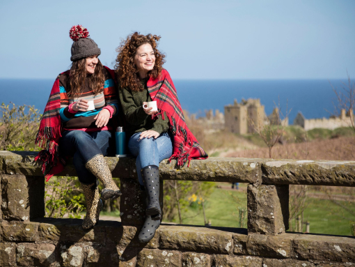 Two women with hot drinks, sitting on a stone wall near a castle