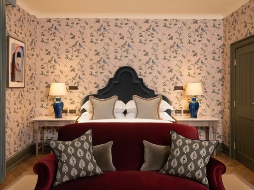 Plush hotel bedroom with red velvet sofa at the foot of a bed with flowery wallpaper on the walls behind it.