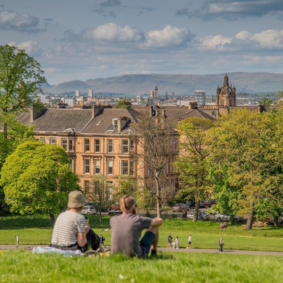 A couple sitting on a hill in park, with Glasgow skyline, and people cycling and walking along park paths.