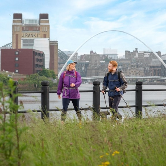 A woman and a man walk along a riverside with iconic buildings and a bridge beyond