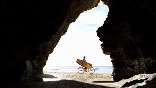 A woman cycles along a beach with her surfboard, silhouetted by a cave entrance.