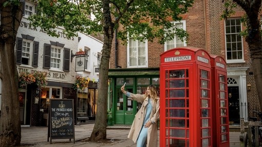 A woman leans against a red telephone box and takes a selfie.