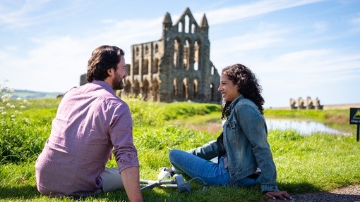 Man and woman sitting on grass with crutches beside them, Whitby Abbey in the background