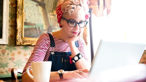 Close up of a blond woman wearing a stripy top and red head scarf, having a coffee and looking down at a lap top screen, smiling.