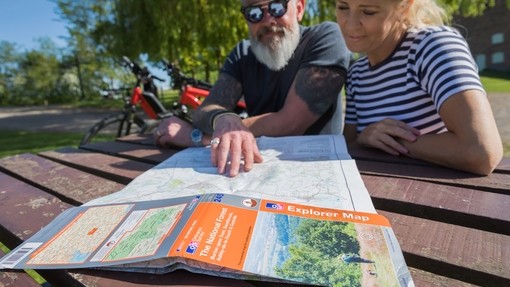 Two people sat at a picnic table, looking at an Ordinance Survey Explorer map of the National Forest. Two Ebikes are parked behind them. National Forest EBike Holidays - Silver award winner for the Ethical, Responsible and Sustainable Tourism Award at the VisitEngland Awards for Excellence 2023.