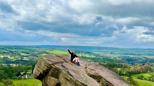 A seated woman waving from a hanging stone, overlooking a country panorama.