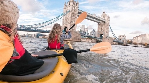 Two women kayaking on a city river towards a bridge.