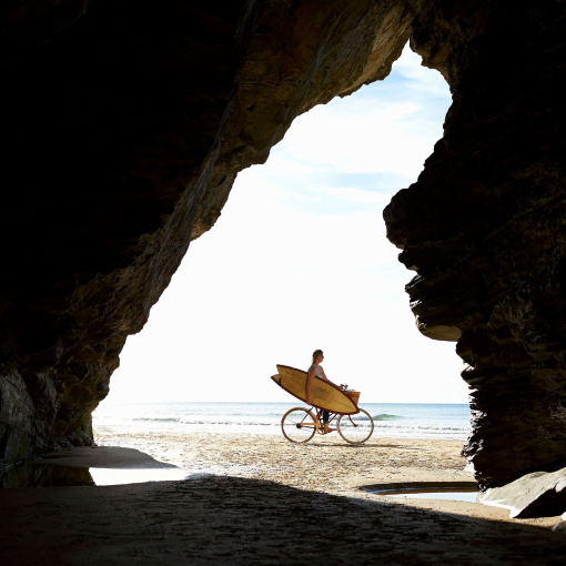A woman cycles along a beach with her surfboard, silhouetted by a cave entrance.