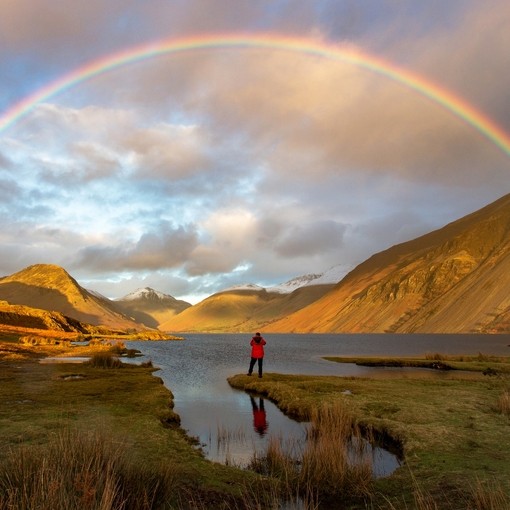 Person standing near lake watching a rainbow in cloudy sky.