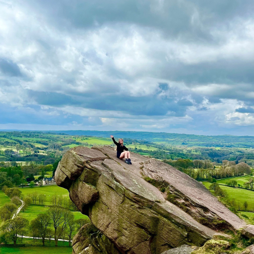 A seated woman waving from a hanging stone, overlooking a country panorama.