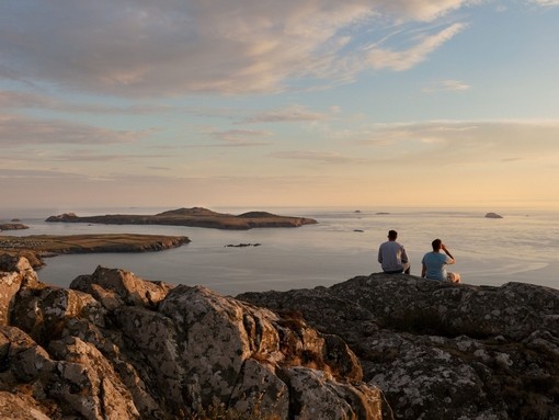 Two men sat on a rock cliff looking out to sea at dusk