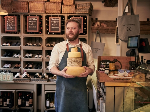 Man wearing blue apron holding a stack of cheeses in a cheese and wine shop