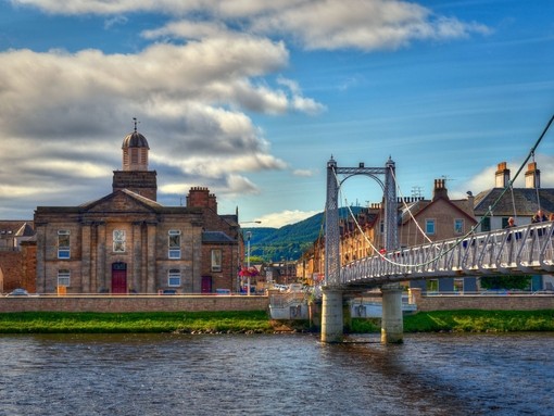 A suspension bridge with people walking across spanning a wide river with houses on the other side