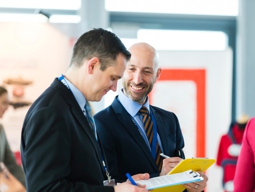 Two men wearing dark suits standing in a professional conference setting smiling and holding papers.