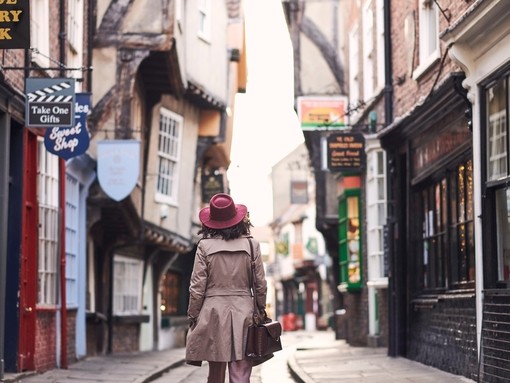 Woman wearing trench coat and pink hat walking through narrow historic street of York, North Yorkshire, England.