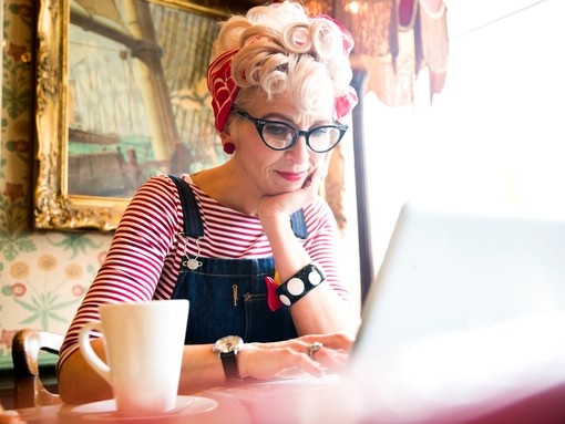 Close up of a blond woman wearing a stripy top and red head scarf, having a coffee and looking down at a lap top screen, smiling.