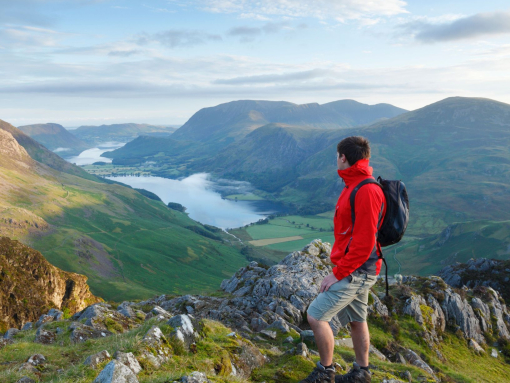 A man wearing a rucksack stands on a mountain fell overlooking hills and a lake