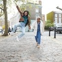 Happy young woman jumping while holding girlfriend's hand walking on a footpath in London street